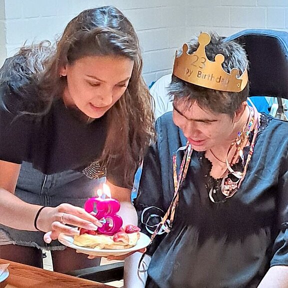 Eine Teilnehmerin feiert ihren Geburtstag. Sie trägt eine goldene Papierkrone auf dem Kopf und bekommt Stück Kuchen mit einer brennenden Kerze überreicht. 